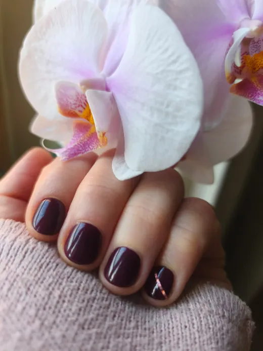 Ongles vernis prune posés par Nathalie, main posée près d'une orchidée blanche